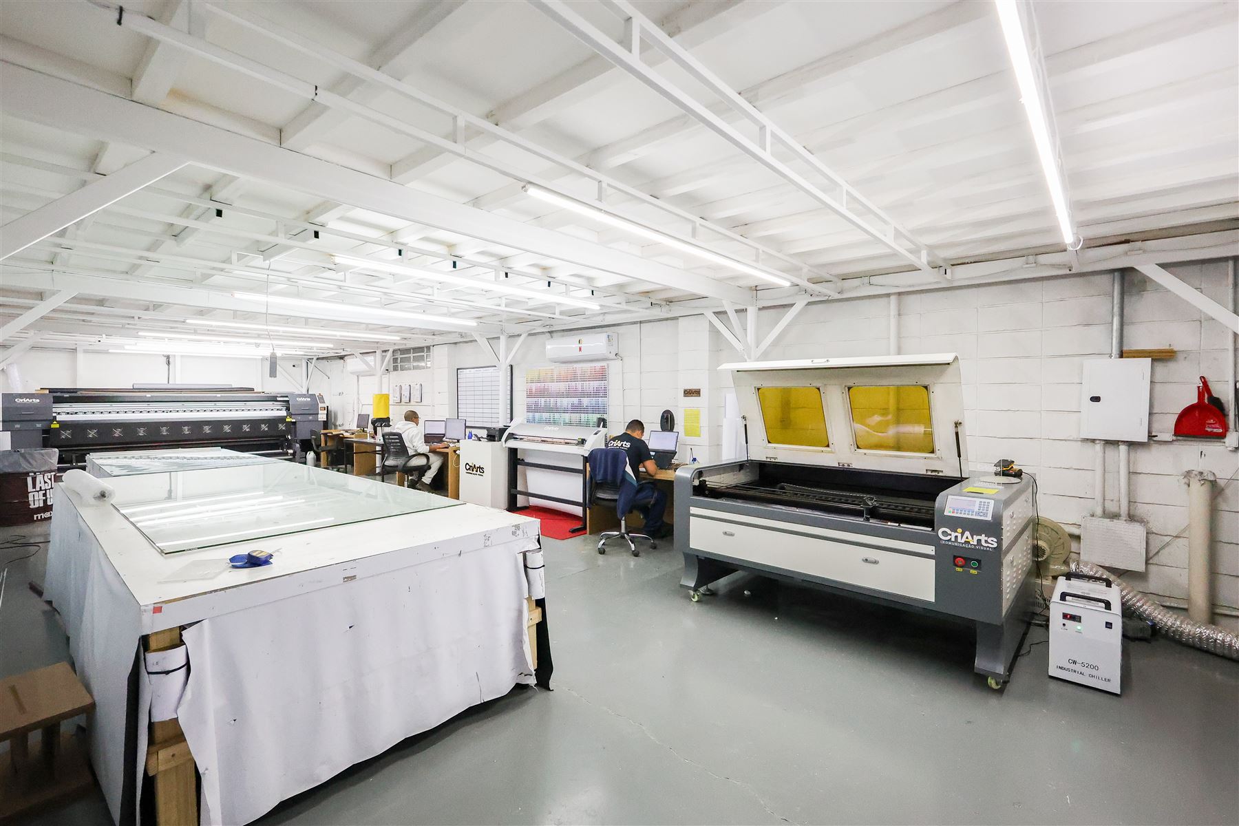 Equipe CriArts trabalhando no chão de fábrica, com impressora e laser ao fundo